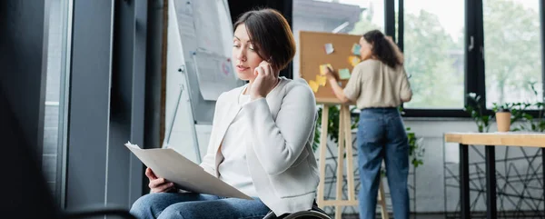 Mujer de negocios con discapacidad mirando documentos y hablando en el teléfono inteligente cerca borrosa colega afroamericano, pancarta - foto de stock