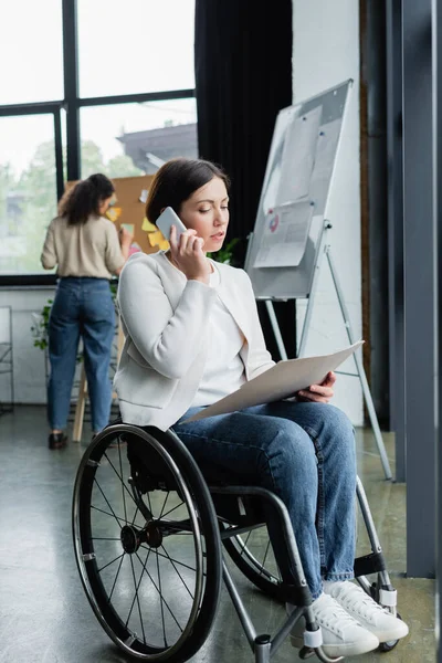 Donna d'affari in sedia a rotelle guardando i documenti e parlando su smartphone vicino sfocato collega afroamericano — Foto stock