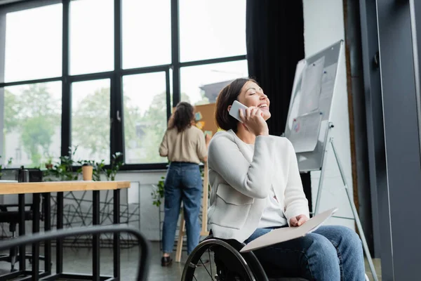 Happy businesswoman in wheelchair talking on smartphone near blurred african american colleague in office — Stock Photo