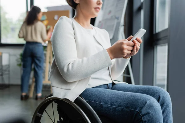 Vista ritagliata di donna d'affari in sedia a rotelle messaggistica sul telefono cellulare vicino collega americano africano su sfondo sfocato — Foto stock