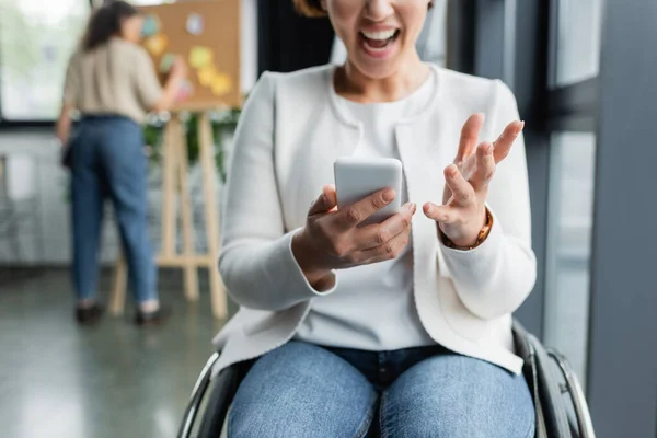 Angry businesswoman in wheelchair using smartphone near african american colleague working on blurred background — Stock Photo