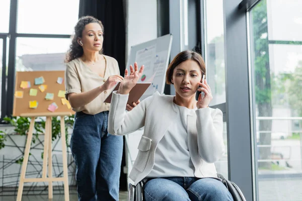 Femme d'affaires en fauteuil roulant parlant sur téléphone portable et montrant geste d'attente à un collègue afro-américain — Photo de stock