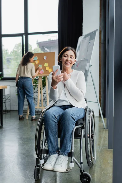 Empresária alegre em cadeira de rodas olhando para o celular perto de colega afro-americano em fundo borrado — Fotografia de Stock
