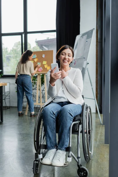 Femme d'affaires joyeuse en fauteuil roulant regardant le téléphone portable près d'un collègue afro-américain sur fond flou — Photo de stock