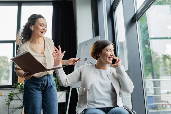 Alegre mujer de negocios en silla de ruedas hablando por teléfono móvil y mostrando gesto de espera a sonriente colega afroamericano - foto de stock