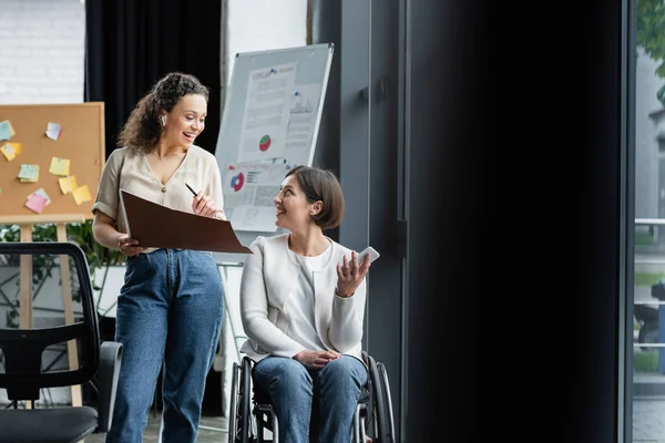 Happy businesswoman in wheelchair talking to african american colleague near blurred flip chart in office — Stock Photo