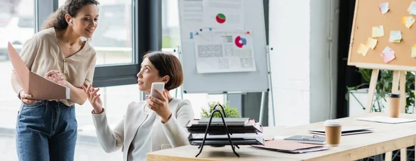 Businesswoman showing smartphone to smiling african american colleague with folder, banner — Stock Photo