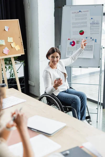 Femme d'affaires souriante en fauteuil roulant tenant smartphone tout en pointant vers tableau à feuilles avec infographies — Photo de stock