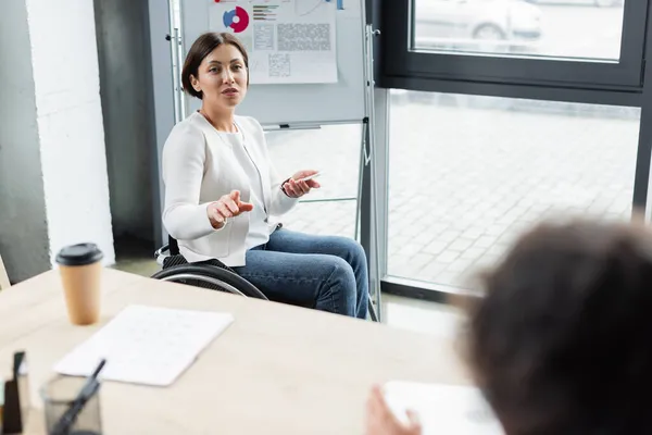 Businesswoman in wheelchair pointing with finger near blurred colleague in office — Stock Photo