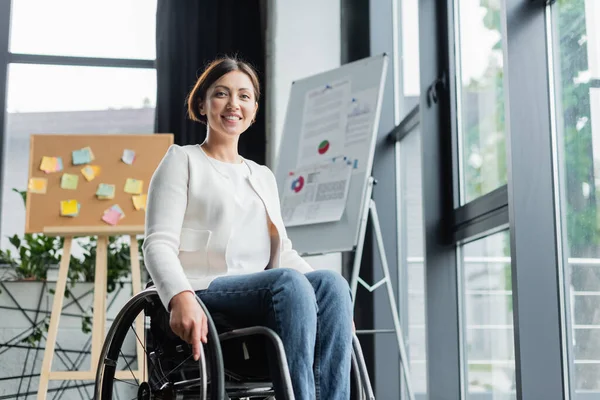 Весела бізнес-леді в інвалідному візку дивиться на камеру біля розмитої інфографіки в офісі — стокове фото