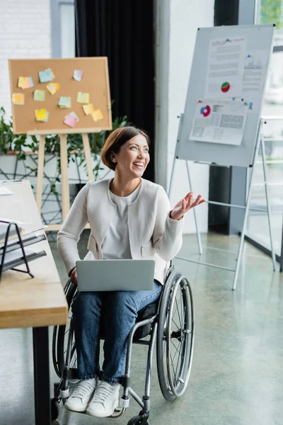 Happy businesswoman with disability pointing with hand while working on laptop near blurred graphs on flip chart — Stock Photo