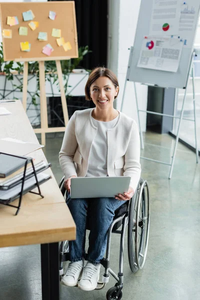 Smiling businesswoman in wheelchair working on laptop near blurred corkboard and flip chart with infographics — Stock Photo