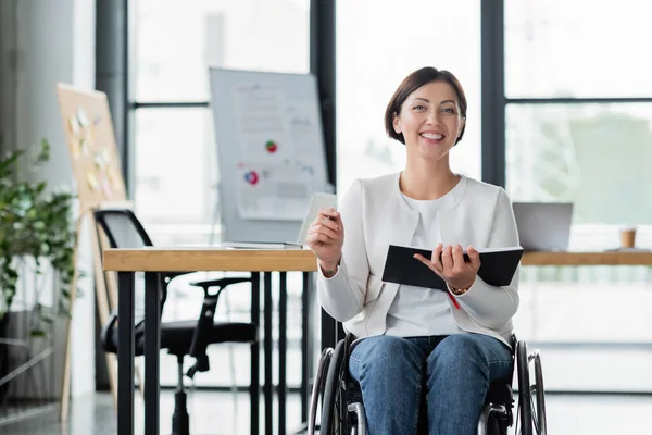 Бізнес-леді в інвалідному візку посміхається на камеру, тримаючи блокнот в офісі — стокове фото