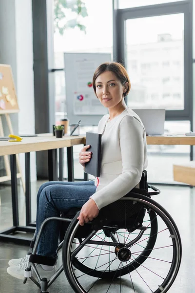Улыбающаяся деловая женщина с ограниченными физическими возможностями держит ноутбук в офисе — стоковое фото