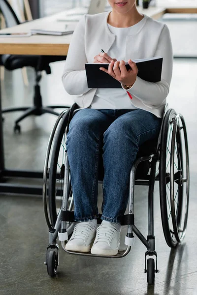 Vista parcial de la mujer de negocios en silla de ruedas escribiendo en un cuaderno mientras trabaja en la oficina - foto de stock
