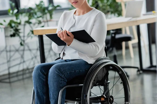 Vue recadrée de femme d'affaires en fauteuil roulant écrit dans un cahier au bureau — Photo de stock