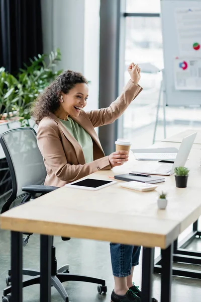 African american businesswoman with takeaway drink showing triumph gesture near laptop in office — Stock Photo