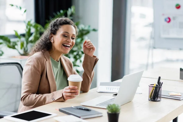 Африканский американский бизнесмен с кофе идти улыбаясь во время видеозвонка на ноутбуке в офисе — стоковое фото