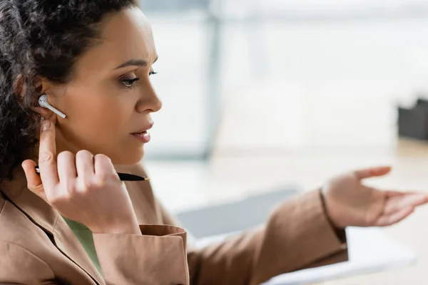 Mujer de negocios afroamericana en auriculares inalámbricos sosteniendo pluma y señalando con la mano en la oficina - foto de stock