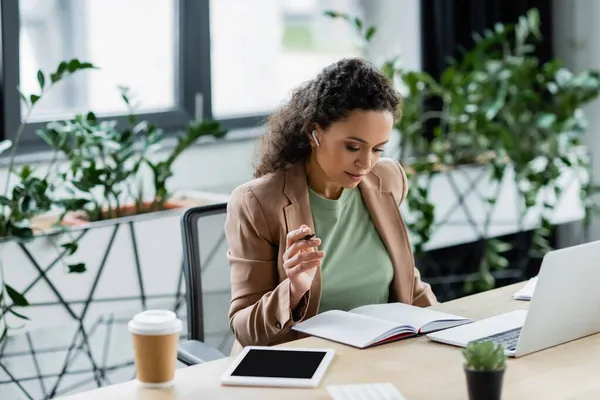 African american businesswoman in wireless headphone looking in notebook near laptop and digital tablet — Stock Photo