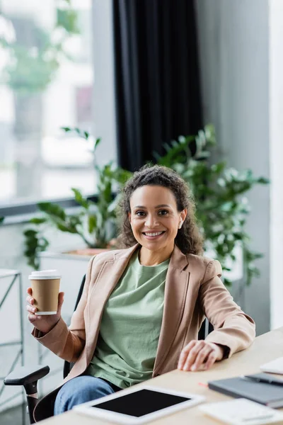Exitosa mujer de negocios afroamericana con café para ir sonriendo a la cámara mientras está sentado en el lugar de trabajo - foto de stock