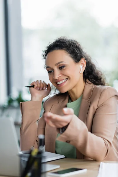 Alegre mujer de negocios afroamericana señalando con la mano durante la videoconferencia en la computadora portátil borrosa - foto de stock