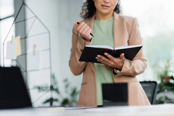 Ausgeschnittene Ansicht einer afrikanisch-amerikanischen Geschäftsfrau, die mit Notizbuch in der Nähe eines verschwommenen Laptops im Büro steht — Stockfoto