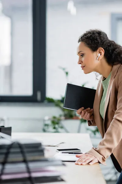 Vista laterale della donna d'affari afroamericana in auricolare senza fili in piedi con notebook sul posto di lavoro in ufficio — Foto stock