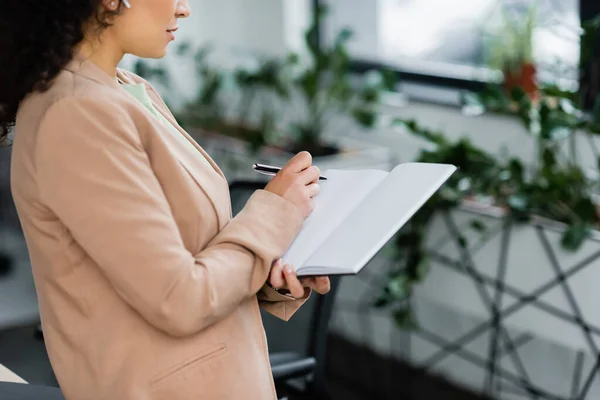 Vista recortada de la mujer de negocios afroamericana escribiendo en cuaderno en blanco en la oficina - foto de stock