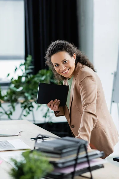 African american businesswoman holding notebook and smiling at camera at workplace in office — Stock Photo