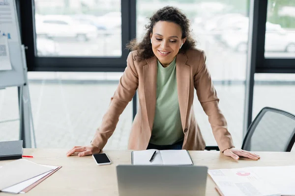 Smiling african american businesswoman standing at work desk during video conference on blurred laptop — Stock Photo