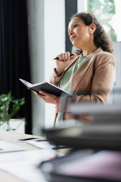 Pensativa mujer de negocios afroamericana de pie con cuaderno y pluma en el lugar de trabajo en la oficina - foto de stock