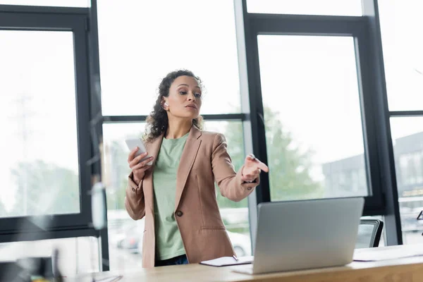 African american businesswoman holding smartphone and pointing at laptop during video call in office — Stock Photo