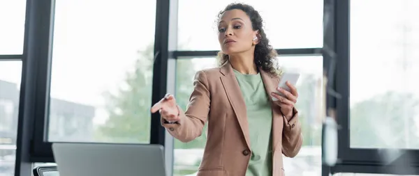 Афроамериканська бізнес-леді в навушниках тримає смартфон і вказує на розмитий ноутбук в офісі, банер — стокове фото