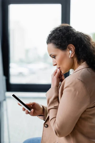 Side view of pensive african american businesswoman in wireless earphone thinking during video call on smartphone — Stock Photo