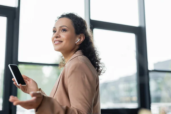 Sorridente mulher de negócios afro-americana em fone de ouvido sem fio apontando com a mão enquanto conversa no smartphone — Fotografia de Stock