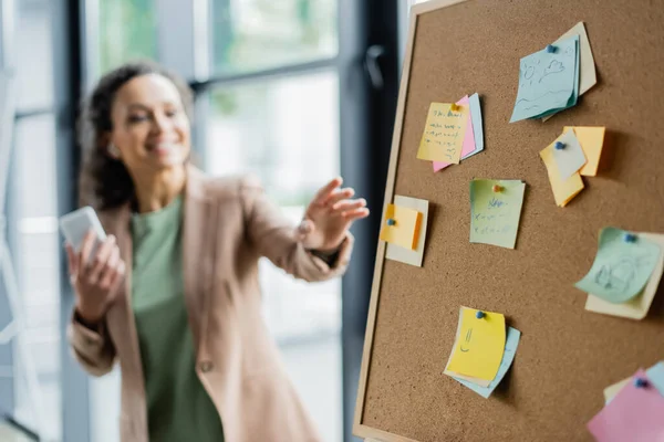 Mujer de negocios afroamericana borrosa con teléfono inteligente apuntando al corcho con notas de papel - foto de stock