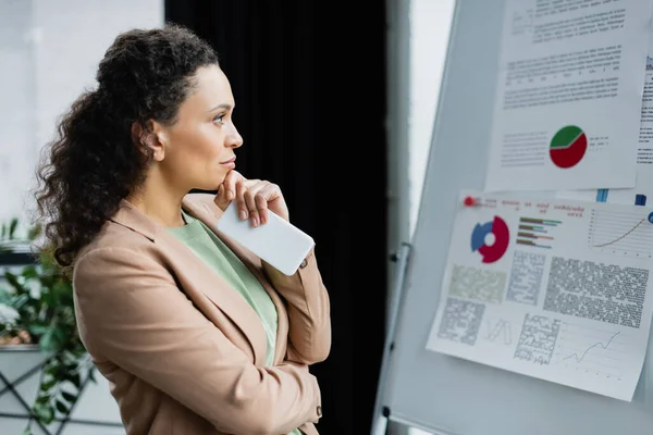 Femme d'affaires afro-américaine réfléchie avec téléphone mobile regardant flip chart avec l'analyse dans le bureau — Photo de stock