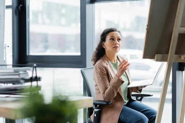 Mujer de negocios afroamericana sentada con el ordenador portátil y señalando el tablero de notas en la oficina - foto de stock