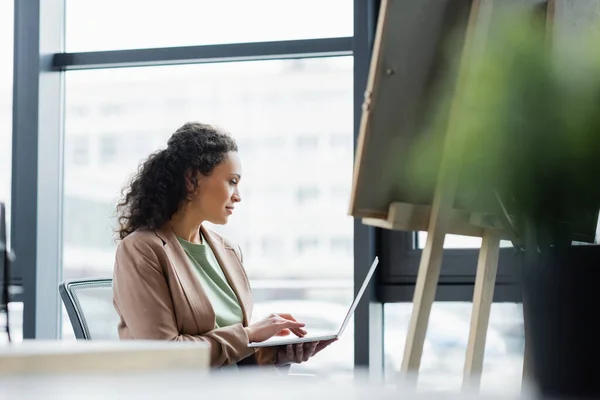 Afro-américaine femme d'affaires en utilisant un ordinateur portable près du tableau de note sur le premier plan flou au bureau — Photo de stock