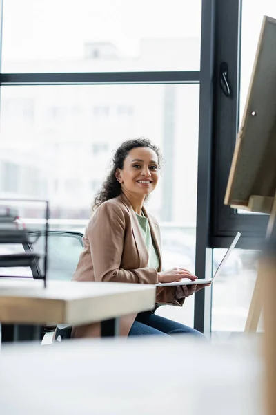 Donna d'affari afro-americana di successo sorridente alla fotocamera durante l'utilizzo del computer portatile in ufficio — Foto stock