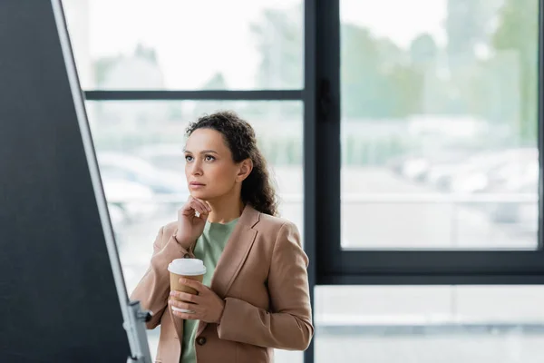 Femme d'affaires afro-américaine réfléchie avec du café pour aller regarder tableau à feuilles brouillées dans le bureau — Photo de stock