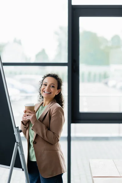 Allegra donna d'affari africana americana con caffè per andare a sorridere alla macchina fotografica vicino flip chart in ufficio — Foto stock