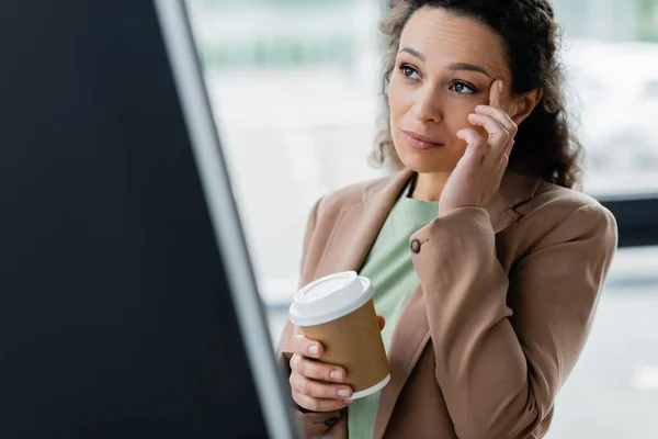 Pensive african american businesswoman with takeaway drink touching head while working on blurred foreground — Stock Photo