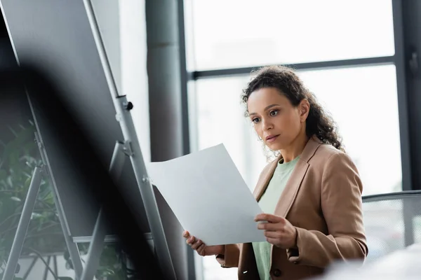 Afro-américaine femme d'affaires regardant le papier tout en travaillant près de tableau à feuilles brouillées dans le bureau — Photo de stock