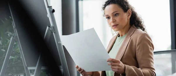 Femme d'affaires afro-américaine regardant le papier tout en se tenant près de flip chart sur le premier plan flou, bannière — Photo de stock