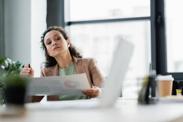 Afro-américaine femme d'affaires regardant ordinateur portable flou tout en travaillant avec des infographies au bureau — Photo de stock