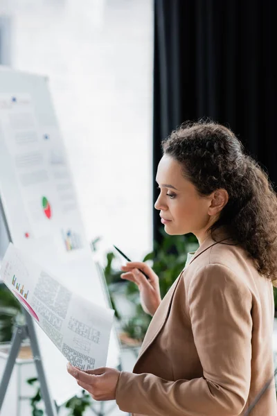 Mulher de negócios afro-americana olhando para infográficos enquanto trabalhava no escritório — Fotografia de Stock