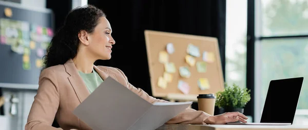 Smiling african american businesswoman holding documents while working on laptop in office, banner — Stock Photo