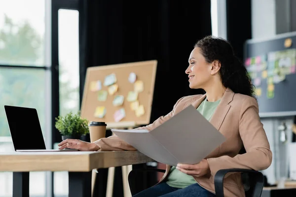 Glücklich afrikanisch-amerikanische Geschäftsfrau arbeitet mit Dokumenten und Laptop mit leerem Bildschirm im Büro — Stockfoto